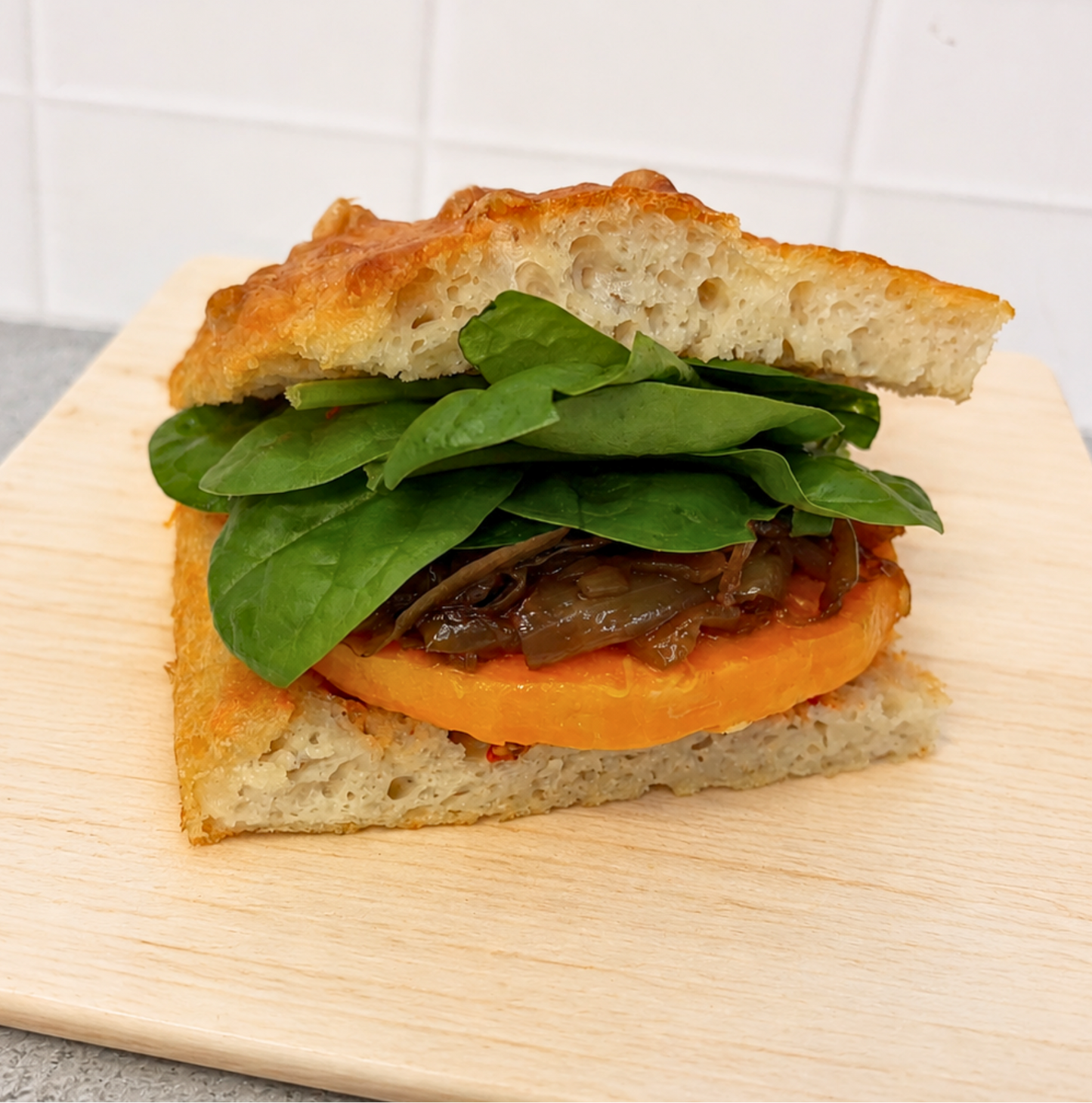 A close-up of a gourmet sandwich on a light wooden board, featuring a ciabatta bread sandwich filled with fresh spinach leaves, caramelized onions, and a thick slice of orange sweet potato, set against a white tiled background.