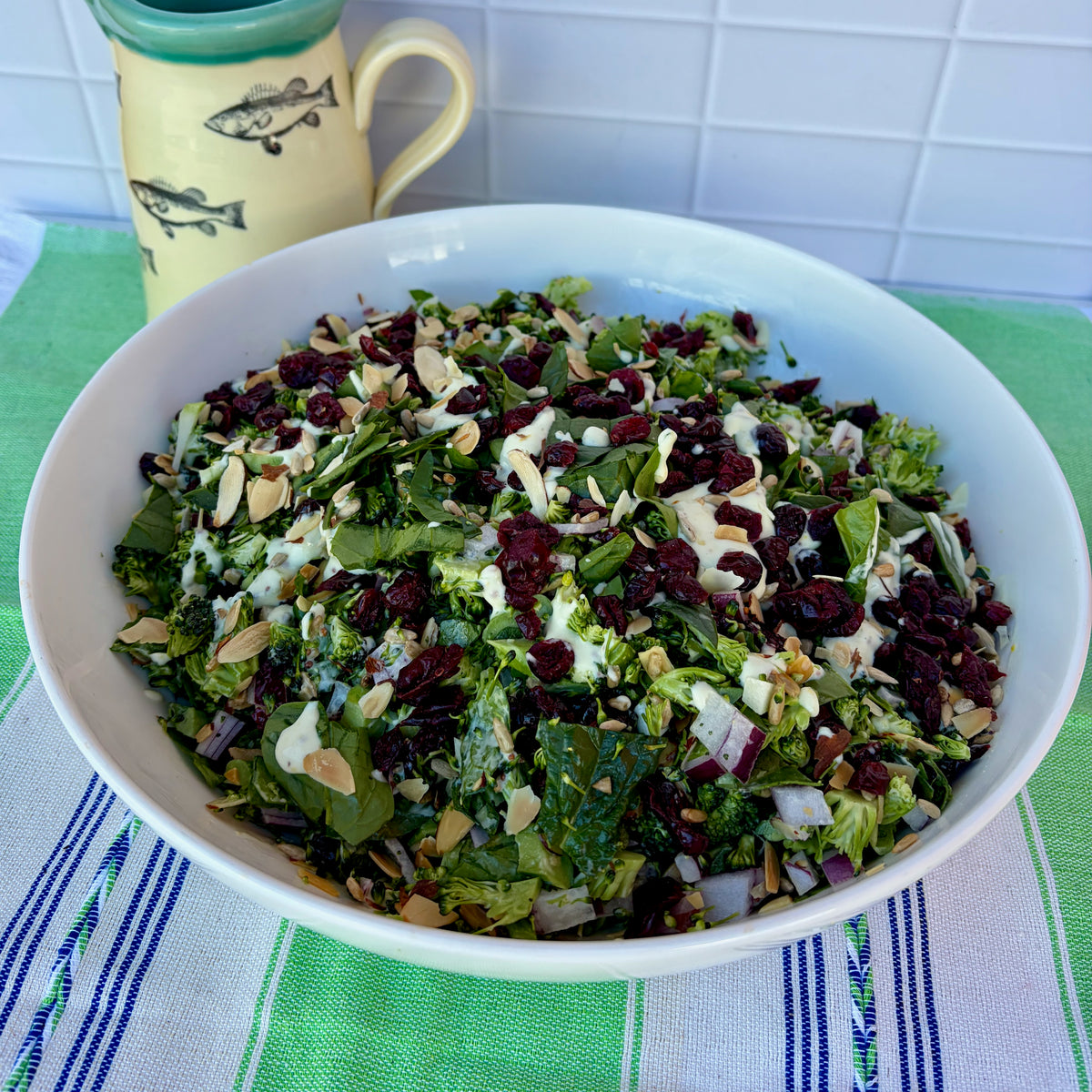 A large white bowl filled with a green kale and broccoli salad topped with dried cranberries, sliced almonds, and a white dressing, set on a green and white striped kitchen towel with a yellow fish-patterned mug in the background.