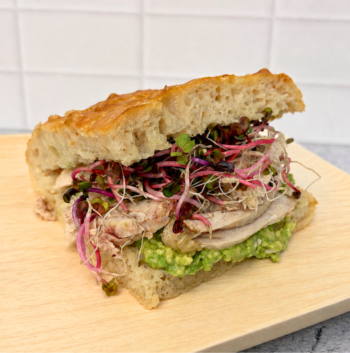 A close-up of a gourmet sandwich on a wooden board, featuring a crusty ciabatta bread, sliced turkey, green avocado spread, and red/purple sprouts, set against a white tiled background.