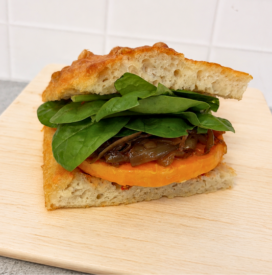 A close-up of a gourmet sandwich on a light wooden board, featuring a ciabatta bread sandwich filled with fresh spinach leaves, caramelized onions, and a thick slice of orange sweet potato, set against a white tiled background.