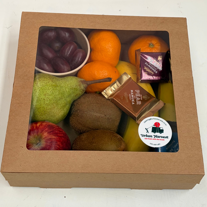 A square brown cardboard gift box with a clear window displaying a fruit assortment including oranges, pears, kiwis, apples, grapes, and packaged snacks, set on a white surface.