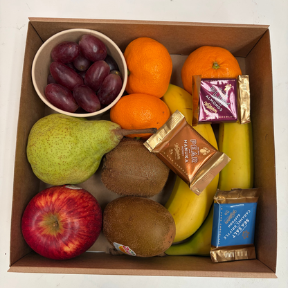 A top-down view of a cardboard gift box filled with assorted fruits and packaged chocolate bars on a white surface. The fruit includes a green pear, a red apple, two kiwis, a bowl of dark grapes, two oranges, and several bananas.