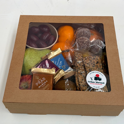 A brown cardboard gift box with a clear window displaying a fruit and snack assortment, including oranges, grapes, a pear, packaged chocolate, and cookies, on a white surface with a pink wall in the background.