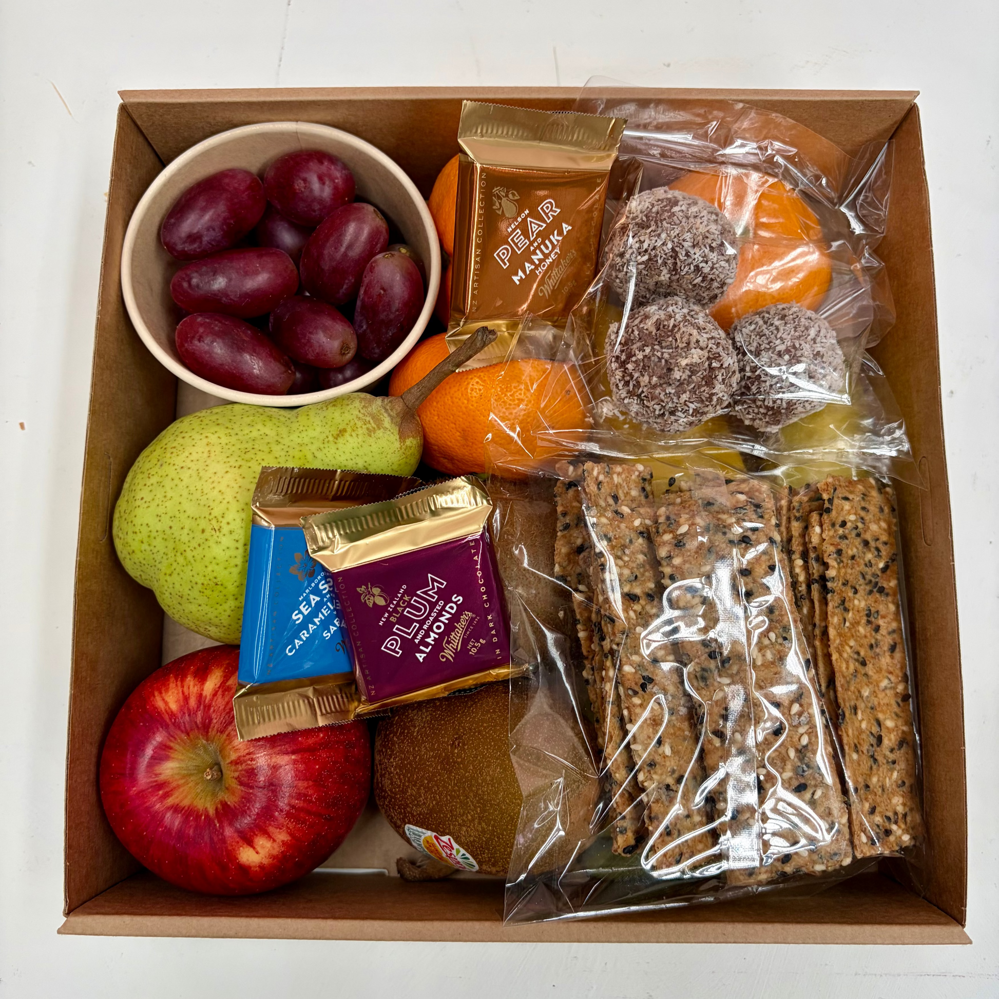 A cardboard gift box filled with assorted snacks and fruit, including a bowl of purple grapes, a green pear, a red apple, a kiwi, packaged chocolate-covered treats, orange slices, and long seeded crackers, photographed from above on a white surface.