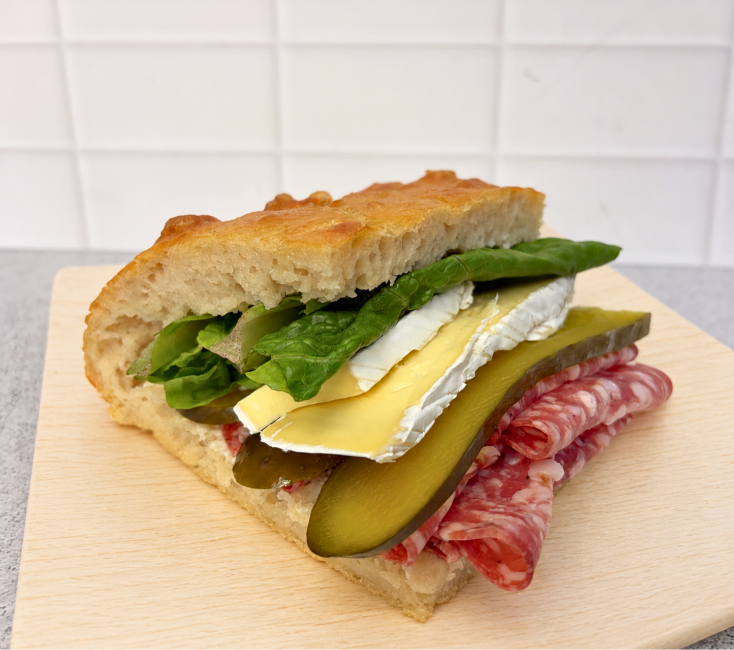 A close-up of a gourmet sandwich on a light wooden board, featuring a rustic bread slice, green lettuce, a slice of white cheese, a pickle slice, and thin slices of cured meat, set against a white tiled background.