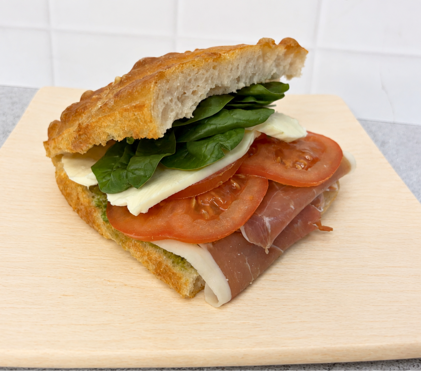 A gourmet sandwich on a light wooden board, featuring a slice of toasted ciabatta bread, fresh green spinach leaves, white cheese, tomato slices, and thin slices of cured prosciutto.