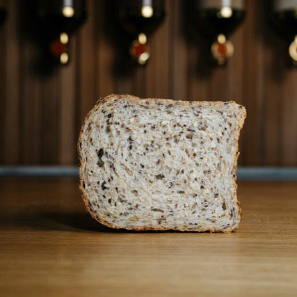 A thick slice of whole wheat bread baked by Wellington sourdough standing upright on a wooden surface, with a blurred background of dark wood paneling and hanging bottles.